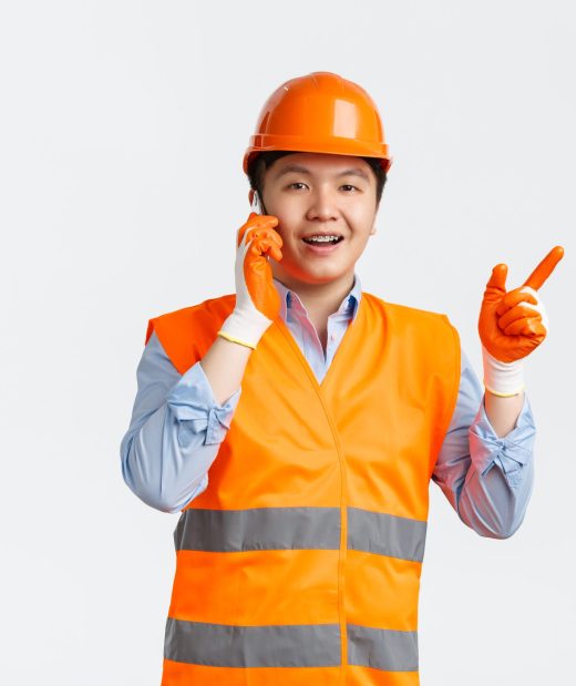 Building sector and industrial workers concept. Smiling asian engineer, construction manager in reflective clothings and helmet pointing finger left while talking on mobile phone, white background.